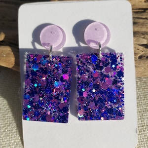 May include: A pair of handmade earrings featuring a rectangular design with a purple glitter finish. The earrings have a small, round, light purple stud at the top and are attached with silver-colored hooks. The earrings are displayed on a white card.