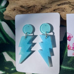 Puede incluir: Un par de pendientes de rayo azul claro. Cada pendiente presenta un perno circular y una forma de rayo, conectados por un pequeño anillo plateado. Los pendientes se exhiben en una tarjeta blanca sobre un fondo de hojas verdes y madera.
