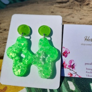 Peut inclure: Boucles d'oreilles en forme de croix vert néon pailleté avec un petit clou rond. Les boucles d'oreilles sont présentées sur une carte blanche à côté d'une carte de visite avec des motifs floraux et du texte.