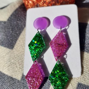 Peut inclure: Une paire de boucles d'oreilles pendantes avec un clou violet et deux pendentifs en forme de losange. Un pendentif est en paillettes vertes, l'autre en paillettes roses. Les boucles d'oreilles sont présentées sur une carte blanche.