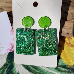 Può includere: Un paio di orecchini verdi glitterati. Ogni orecchino presenta un piccolo perno rotondo verde lime collegato a un ciondolo rettangolare pieno di glitter. I ciondoli sono di un verde vibrante con macchie di vari colori. Gli orecchini sono esposti su una carta bianca.