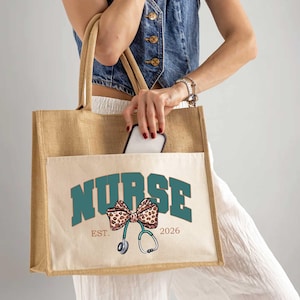 May include: A beige tote bag with a front pocket featuring the word "NURSE" in teal block letters. Below the word is a bow with a leopard print design and a stethoscope. The bag has a "Est. 2026" text.