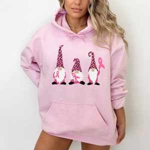 May include: Pink hoodie with a graphic of three gnomes wearing pink and black leopard print hats and holding pink ribbons. The gnomes are standing in a row.