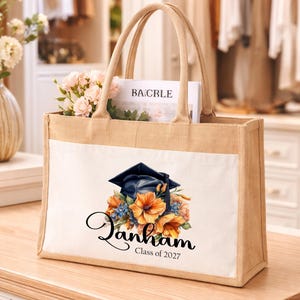 May include: A rectangular jute and canvas tote bag featuring a printed graphic of a dark blue graduation cap resting on a cluster of orange and blue flowers. The text "Janham" in a black script font and "Class of 2027" in a simple font appear below.