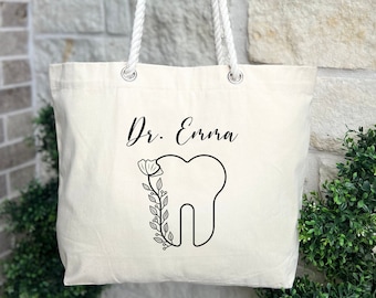 May include: A white canvas tote bag with rope handles. The bag has a black and white design of a tooth with a flower and the text "Dr. Enna".
