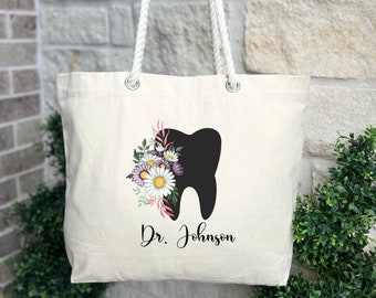 May include: A white canvas tote bag with rope handles. The bag features a black silhouette of a tooth with pink and white flowers. The text "Dr. Johnson" is printed below the tooth.