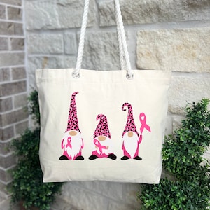 May include: A white canvas tote bag with pink and black leopard print gnomes. The gnomes are holding pink ribbons. The bag has rope handles.