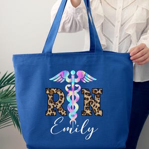 May include: A blue canvas tote bag with a rainbow-colored medical symbol and the letters "RN" in leopard print. The name "Emily" is written below the letters.