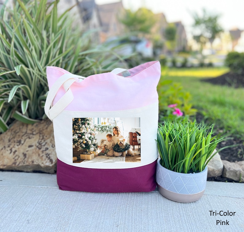 May include: A tri-color tote bag with a pink, white, and burgundy design. The bag features a photo of a family with a dog in front of a Christmas tree.