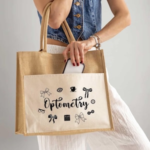May include: A large, rectangular tote bag made of tan burlap with a cream-colored pocket. The pocket features the word "Optometry" in script, surrounded by black graphic icons. A smartphone is partially visible in the pocket.