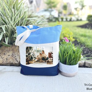 May include: A blue, white, and navy blue tote bag with a photo of a woman and child in front of a Christmas tree. The bag has a white canvas section with the photo and a blue canvas section at the bottom.
