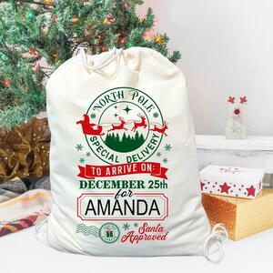 May include: A white canvas Santa sack with a red and green design. The design features Santa Claus in his sleigh with reindeer flying over a green forest. The text reads "North Pole Special Delivery To Arrive On: December 25th for Amanda Santa Approved".