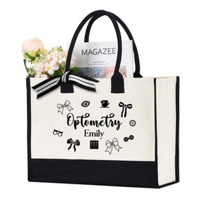 May include: A white tote bag with black handles and a black bottom. The bag features the word "Optometry" and the name "Emily" in black script, along with eye-related icons. The bag contains a magazine and flowers.