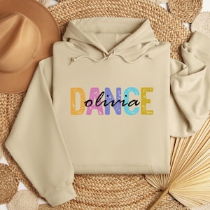 May include: A beige hoodie with the word "DANCE" in colorful letters and the name "olivia" in a script font. The hoodie has a drawstring hood and long sleeves. The background includes a woven mat, a hat, and a dried palm leaf.