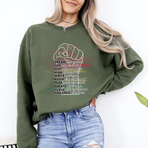 May include: An olive green sweatshirt with a white fist graphic and text. The text includes phrases like "Dream like Martin," "Write like Maya," and "Believe like Thurgood." The sweatshirt is paired with blue jeans.