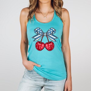 May include: A turquoise tank top featuring a graphic of two red cherries with white star accents, tied together with a blue bow adorned with white stars. The design is centered on the front of the tank top.