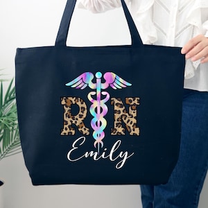 May include: Black canvas tote bag with a colorful tie-dye medical symbol and the letters "RN" in leopard print. The name "Emily" is written below the symbol.