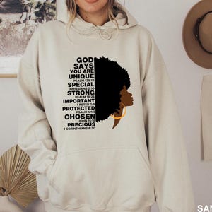 May include: A beige hoodie featuring the text "GOD SAYS YOU ARE UNIQUE" with a silhouette of a woman with an afro and gold hoop earrings. The hoodie also includes Bible verses. The hoodie has a front pocket and a drawstring hood.