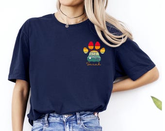 May include: Navy blue t-shirt with a paw print design. The paw print features a stethoscope and a landscape scene. The name "Sarah" is printed below the paw print. The shirt is paired with blue jeans.