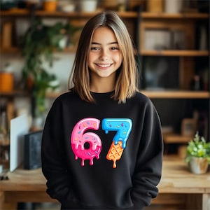 May include: A black sweatshirt featuring the number "67" in a colorful, playful design. The "6" is pink with sprinkles, and the "7" is blue with a melting ice cream cone. The sweatshirt is worn by a smiling person.