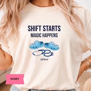 Puede incluir: Camiseta color marfil con el texto azul marino "SHIFT STARTS MAGIC HAPPENS". Debajo, dos zuecos azul claro y un estetoscopio. Las palabras "L&D Nurse" están impresas debajo.