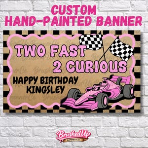 May include: A hand-painted banner with a pink race car, checkered flags, and the text "TWO FAST 2 CURIOUS HAPPY BIRTHDAY KINGSLEY". The banner has a brown background with a pink and black border.