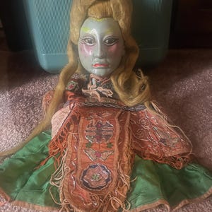 May include: A doll with a pale blue face, painted features, and long blonde hair. The doll wears an orange fabric garment with intricate embroidery, metallic thread detailing, and long fringe, layered over a bright green skirt with a ruffled hem.