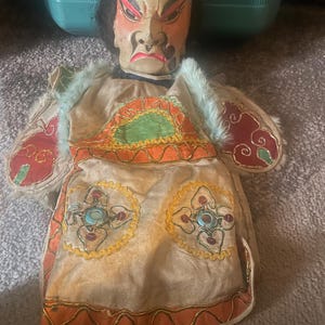 May include: A hand puppet featuring a painted face with dark hair, arched eyebrows, and a stern expression. The puppet wears a beige fabric garment adorned with orange, green, and red embroidered circular patterns, accented by light blue faux fur trim.