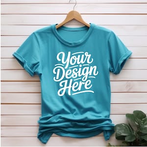K&ouml;nnte beinhalten: Ein t&uuml;rkisfarbenes T-Shirt mit hochgekrempelten &Auml;rmeln, das an einem Holzb&uuml;gel h&auml;ngt. Das Shirt hat den Text "Your Design Here" in Wei&szlig; auf der Vorderseite.
