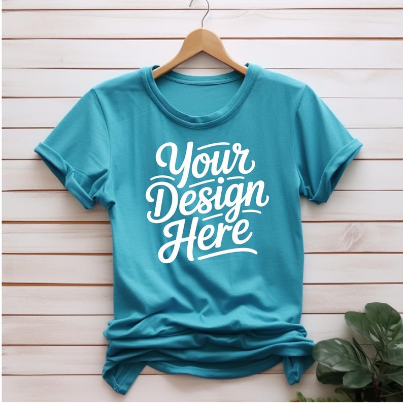 K&ouml;nnte beinhalten: Ein t&uuml;rkisfarbenes T-Shirt mit hochgekrempelten &Auml;rmeln, das an einem Holzb&uuml;gel h&auml;ngt. Das Shirt hat den Text "Your Design Here" in Wei&szlig; auf der Vorderseite.