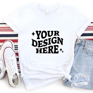 May include: White t-shirt with the text "YOUR DESIGN HERE" in black, paired with light blue jeans and white sneakers. The shirt is knotted at the waist, and the sneakers have red accents. A red, white, and blue striped fabric is in the background.