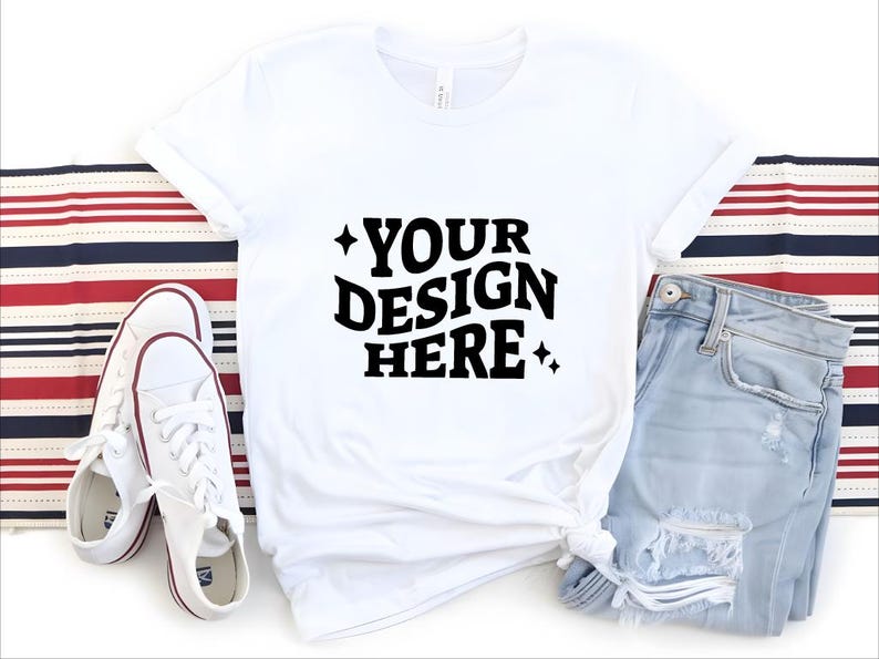 May include: White t-shirt with the text "YOUR DESIGN HERE" in black, paired with light blue jeans and white sneakers. The shirt is knotted at the waist, and the sneakers have red accents. A red, white, and blue striped fabric is in the background.