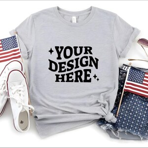 May include: A light gray t-shirt with the text "YOUR DESIGN HERE" in black. The shirt is styled with white sneakers, denim shorts, sunglasses, and two American flags. The overall theme is patriotic and casual.