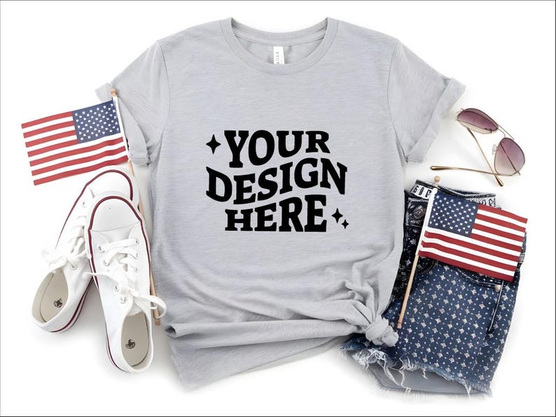 May include: A light gray t-shirt with the text "YOUR DESIGN HERE" in black. The shirt is styled with white sneakers, denim shorts, sunglasses, and two American flags. The overall theme is patriotic and casual.