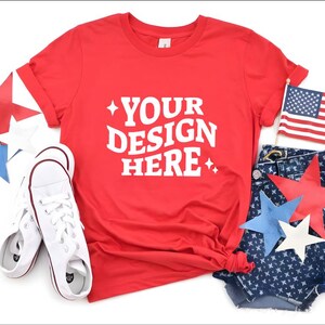 May include: A red t-shirt with the text "YOUR DESIGN HERE" in white, paired with denim shorts, white sneakers, and star-shaped decorations in red, white, and blue. An American flag is also included.