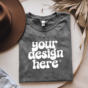 May include: A gray t-shirt with the text "your design here" in white, with a star above the word "here". The t-shirt is folded and laid on a white surface with a brown hat and a necklace.