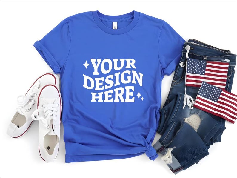 May include: A royal blue t-shirt with the text "YOUR DESIGN HERE" in white, paired with white sneakers, distressed denim jeans, and two American flag patches. The t-shirt is knotted at the bottom.