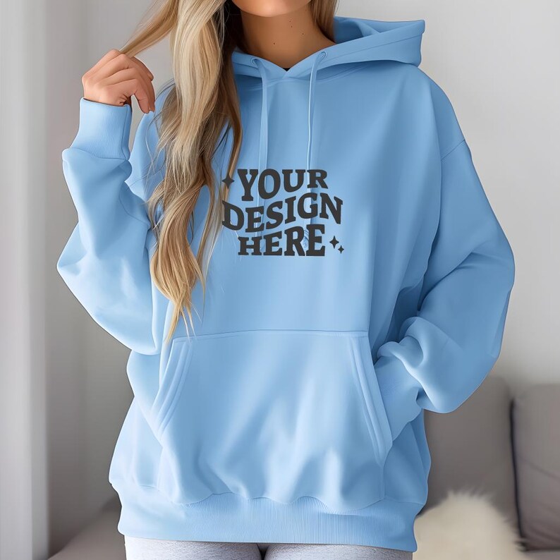 Pu&ograve; includere: Una felpa con cappuccio blu chiaro con una grande tasca sul davanti. La felpa ha il testo "YOUR DESIGN HERE" stampato sul davanti.