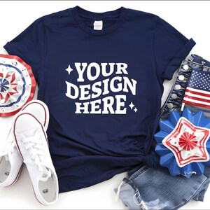 May include: Navy blue t-shirt with the text "YOUR DESIGN HERE", paired with white sneakers, denim shorts, and patriotic decorations. The ensemble includes a red, white, and blue paper fan and a star-shaped pin.