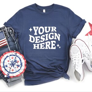 May include: Navy blue t-shirt with the text "YOUR DESIGN HERE" in white, paired with denim jeans, white sneakers, and patriotic decorations including an American flag and star-shaped ornaments.