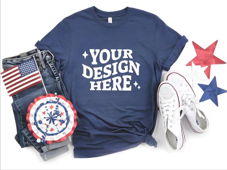 May include: Navy blue t-shirt with the text "YOUR DESIGN HERE" in white, paired with denim jeans, white sneakers, and patriotic decorations including an American flag and star-shaped ornaments.