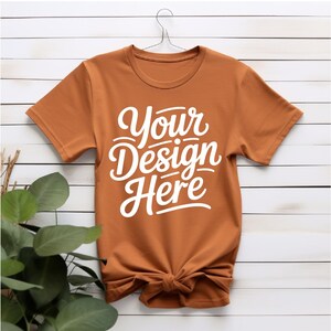 K&ouml;nnte beinhalten: Ein rostfarbenes T-Shirt mit kurzen &Auml;rmeln und einem Rundhalsausschnitt. Das Shirt hat den Text "Your Design Here" in Wei&szlig; auf der Vorderseite aufgedruckt.