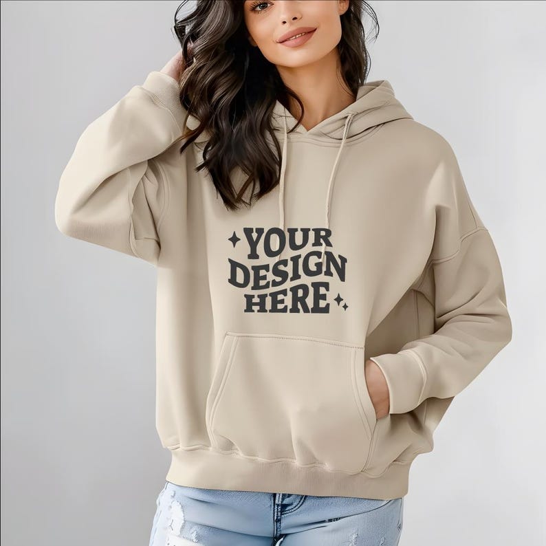 Pu&ograve; includere: Una felpa con cappuccio beige con una grande tasca sul davanti. La felpa ha la scritta "YOUR DESIGN HERE" stampata sul davanti.