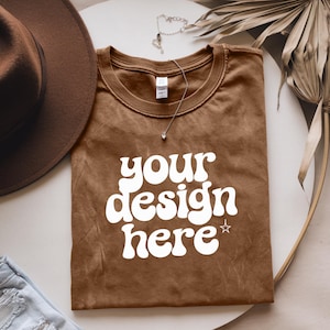 Puede incluir: Una camiseta marrón con el texto "your design here" en blanco, con una estrella sobre la palabra "here". La camiseta está doblada y colocada sobre una superficie blanca.