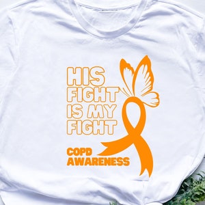 Puede incluir: Camiseta blanca con una cinta naranja y un diseño de mariposa. El texto en la camiseta dice "His Fight Is My Fight COPD Awareness".