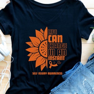 Pode incluir: Camiseta preta com um gráfico de girassol laranja e o texto "Life can change in an instant" com uma fita laranja. O texto "Self Injury Awareness" está abaixo do gráfico.