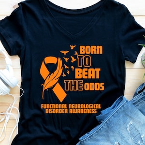 Puede incluir: Camiseta negra con una cinta naranja, una pluma y pájaros volando. El texto dice "Born to Beat the Odds" y "Functional Neurological Disorder Awareness".
