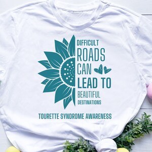 May include: White t-shirt with a teal sunflower graphic and the text "Difficult Roads Can Lead To Beautiful Destinations Tourette Syndrome Awareness".