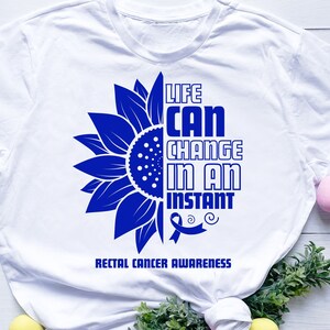 Puede incluir: Camiseta blanca con un gráfico de girasol azul y el texto "Life Can Change In An Instant" con una cinta azul. El texto "Rectal Cancer Awareness" está debajo del gráfico.