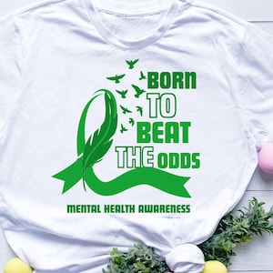 May include: White t-shirt with a green ribbon and a feather design. The text "Born to Beat the Odds" is printed on the shirt. The words "Mental Health Awareness" are printed below the design.